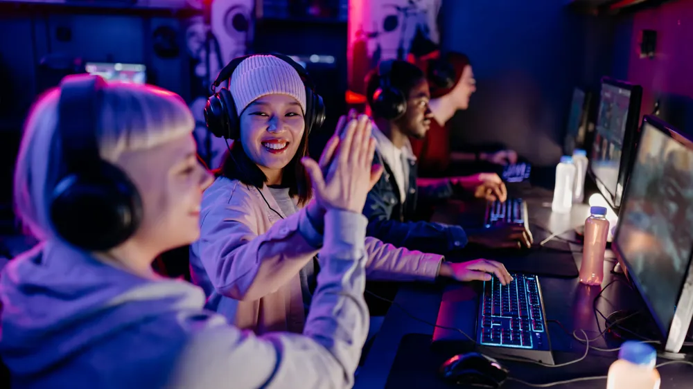 ALT Text :Four people wearing headsets are playing video games on computers in a dimly lit room. Two of them are high-fiving each other.